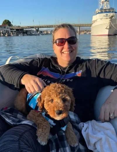 A woman wearing sunglasses and a dark hoodie relaxes in a boat, smiling, while holding a mini red poodle in a blue harness. Boats and a bridge are visible in the background on a sunny day.