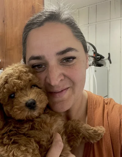 A woman with dark hair pulled back, wearing an orange shirt, smiles while holding a fluffy brown mini poodle puppy close to her face in a cozy indoor setting.