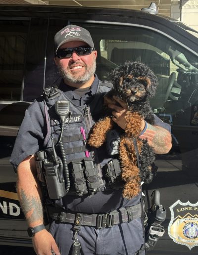 A smiling police officer in uniform holds a small black and brown puppy in front of a police vehicle. The officer, wearing sunglasses and a cap, proudly cradles one of the adorable poodle puppies Utah residents love.